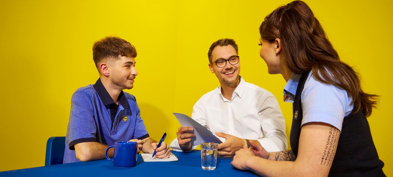 Zwei Lidl-Mitarbeitende und ein Bewerber im Hemd tauschen sich in entspannter Atmosphäre während eines Gesprächs an einem blauen Tisch aus.