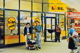 Alte Aufnahme einer Lidl-Filiale mit Kunden, Einkaufswagen und dem Werbespruch "Lidl ist billig" an der Fensterscheibe.