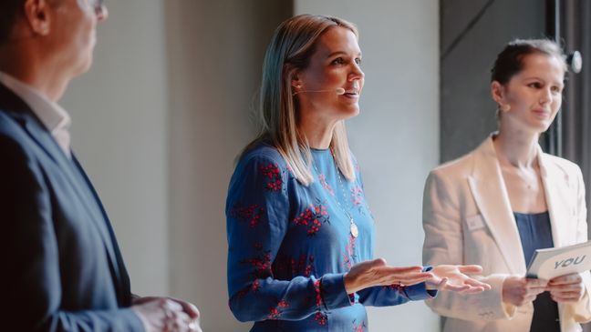 Fokus auf eine engagiert sprechende Sprecherin in Blau, die mit zwei Personen im Rahmen der Mitarbeiter Initiative YOU auf der Bühne steht.