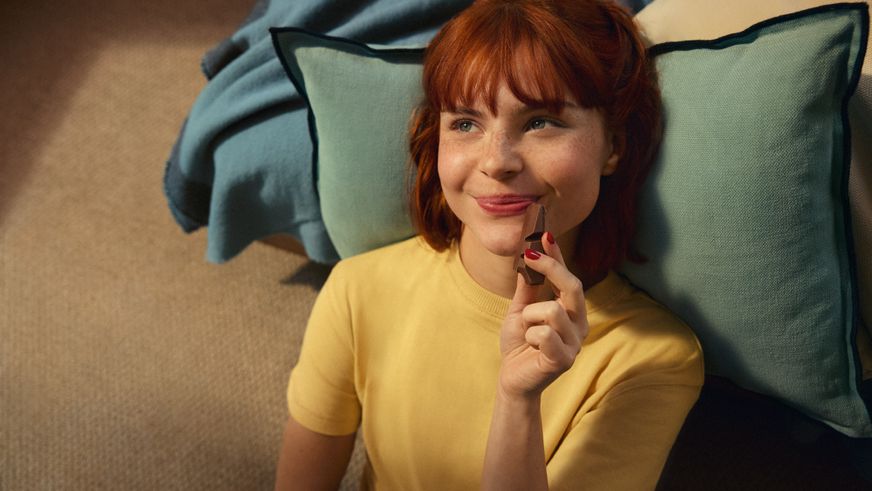 Eine lächelnde junge Frau genießt entspannt ein Stück Schokolade und blickt dabei zufrieden zur Seite.