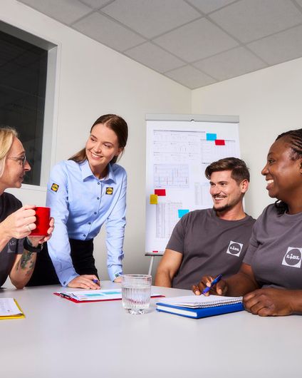 Vier diverse Lidl-Mitarbeitende in Dienstkleidung tauschen sich positiv und kooperativ bei einem Meeting aus.
