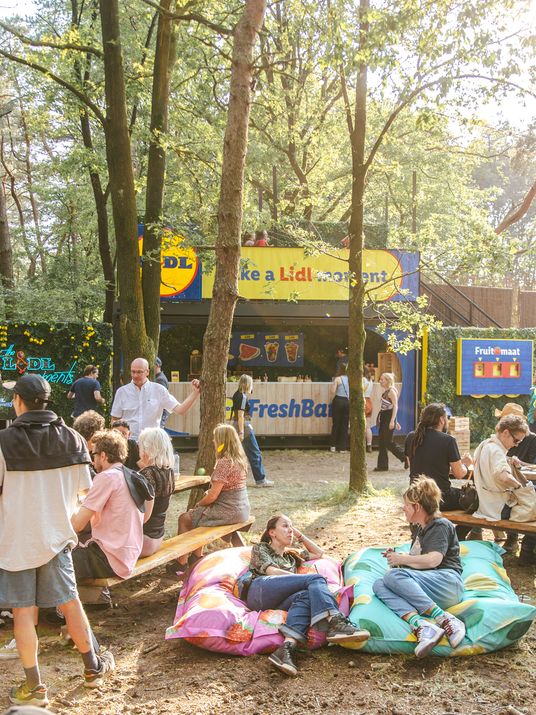 Menschen entspannen auf Bierbänken und Sitzkissen in der Sonne im Wald an einem Lidl Food-Stand und verbingen gemeinsame Zeit.