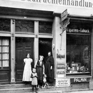 Arhivska crno-bijela fotografija originalne trgovine A. Lidl & Cie. s članovima obitelji i psom ispred ulaza.