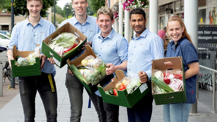 Fünf lächelnde Mitarbeiter präsentieren vor einer Filiale grüne Kisten voller Lebensmittelspenden.