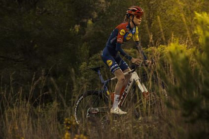 Eine Radsportlerin des Lidl-Trek Teams fährt in Teambekleidung auf einem Rennrad, umgeben von hoher, trockener Vegetation vor einem dunklen Waldhintergrund.