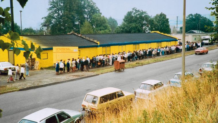 Aufnahme einer langen Menschenschlange vor einer gelben Verkaufshalle mit DDR-Autos im Vordergrund.