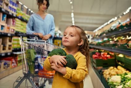 Ein kleines Mädchen trägt entschlossen zwei Köpfe Brokkoli im Supermarkt, während ihre Mutter den Einkaufswagen schiebt.