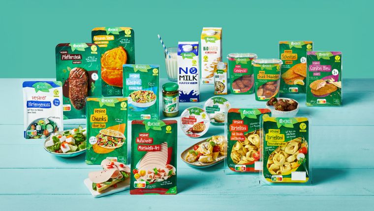 Eine bunte Auswahl an veganen Vemondo-Produkten präsentiert sich ansprechend auf einem hellblauen Holztisch vor mintgrünem Grund.