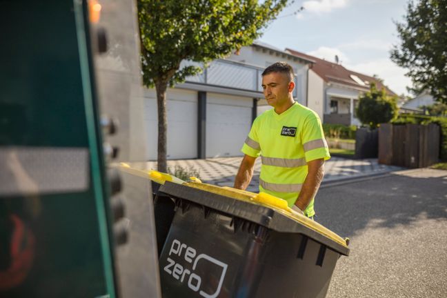 Ein Mitarbeiter in Warnkleidung schiebt eine PreZero-Wertstofftonne zur Leerung durch ein Wohngebiet.