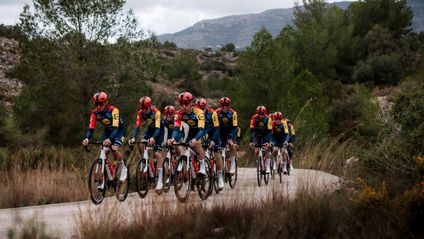 Ein Team von Lidl-Trek Profi-Radsportlern fährt in ihrer rot-gelb-blauen Teambekleidung in einer Formation auf einer kurvigen, ländlichen Straße. Im Vordergrund sind Fahrer in dichten Reihen zu sehen, gefolgt von weiteren Fahrern. Der Hintergrund zeigt eine trockene, bewachsene Hügellandschaft unter einem bewölkten Himmel.