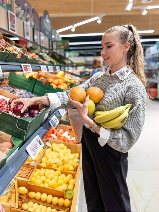 Junge Frau greift lächelnd nach dem frischen Obst im Regal und trägt es in ihrem Arm.