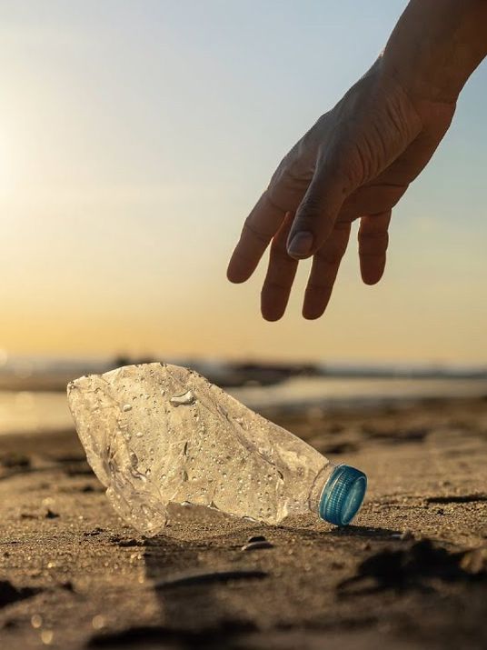 Eine Hand greift nach einer leeren Plastikflasche am Strand im Sonnenuntergang zur Unterstützung von Plastic Free Greece.
