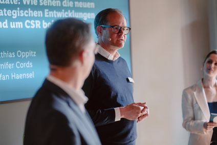 Sprecher mit Brille moderiert eine Diskussionsrunde der Mitarbeiter Initiative YOU vor einer blauen Infografik auf Leinwand.
