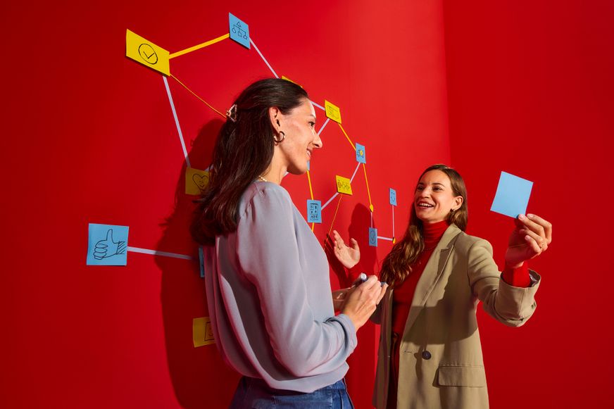 Zwei Lidl Kolleginnen planen lächelnd eine kreative Strategie mit bunten Notizzetteln an einer leuchtend roten Wand.