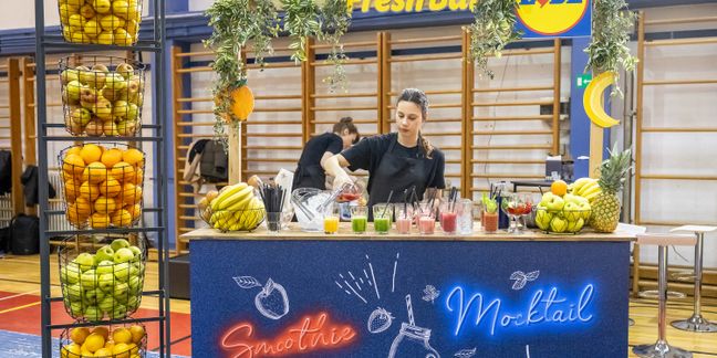 Opcija 1: Preporučena (kratka i opisna)  „Djevojka priprema voćne napitke za brendiranim Lidlovim 'Fresh Bar' štandom uz košare punim svježeg voća.
