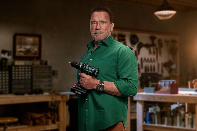 Arnold Schwarzenegger hält eine Parkside Akku-Bohrmaschine entschlossen in einer Werkstatt-Umgebung.