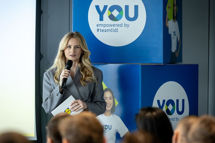 Govornica s mikrofonom i Lidlovom bilježnicom obraća se publici ispred vizuala 'YOU empowered by #teamlidl'.
