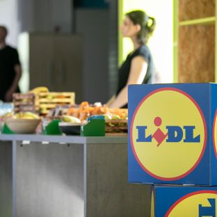 Kutija s Lidlovim logotipom u prvom planu, dok je u pozadini stol sa svježim voćem i vodom te dvoje ljudi