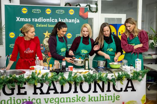 Sudionice radionice kuhanja u zelenim pregačama zajedno pripremaju veganska jela za kuhinjskim stolom.