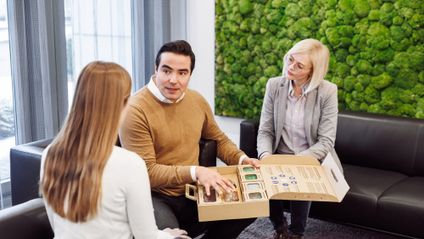 Ein Berater präsentiert zwei Kundinnen in einem modernen Büro eine Musterbox mit nachhaltigen Verpackungsmaterialien.