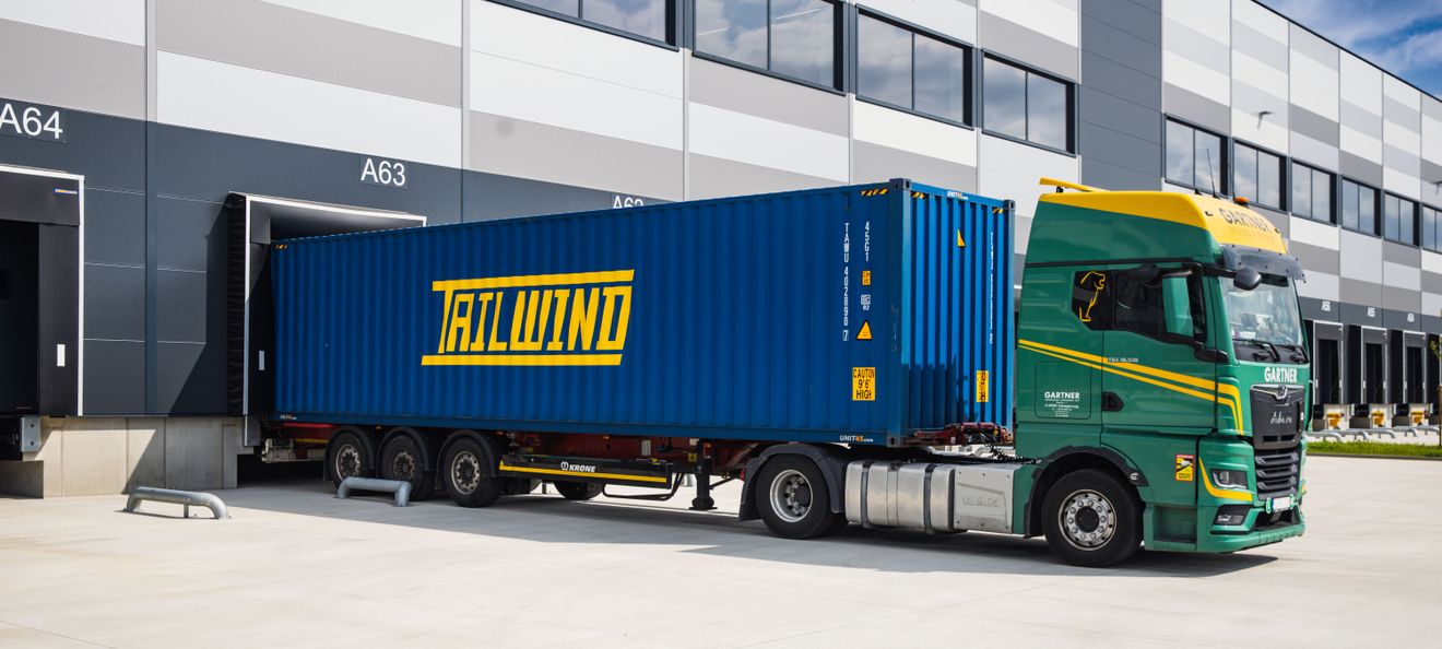 Ein grüner MAN-Lkw verlädt einen blauen Tailwind Container an der Logistikrampe eines modernen Warenlagers.