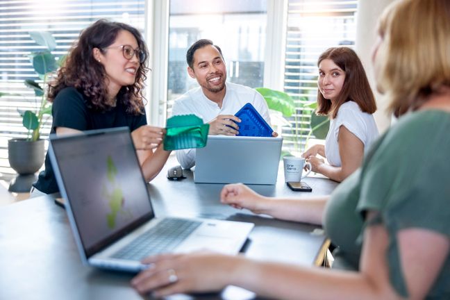 Ein Team begutachtet und bespricht lächelnd verschiedene Prototypen für nachhaltige Kunststoffverpackungen im Büro.