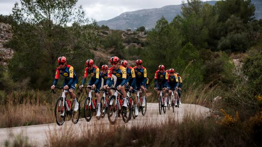 A group of Lidl-Trek professional cyclists in their red, yellow, and blue team kit, training in formation on a rural road in a hilly landscape. In the foreground, the riders are arranged in two rows, with the lead riders clearly visible. The background shows dry vegetation, pine trees, and mountains under an overcast sky.