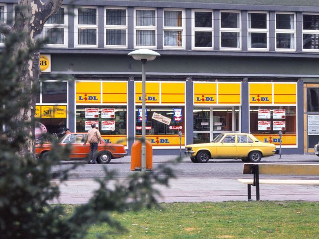 Eine historische Lidl-Filiale mit gelben Fensterfronten und zwei geparkten Oldtimern versprüht den Charme der 1970er-Jahre.