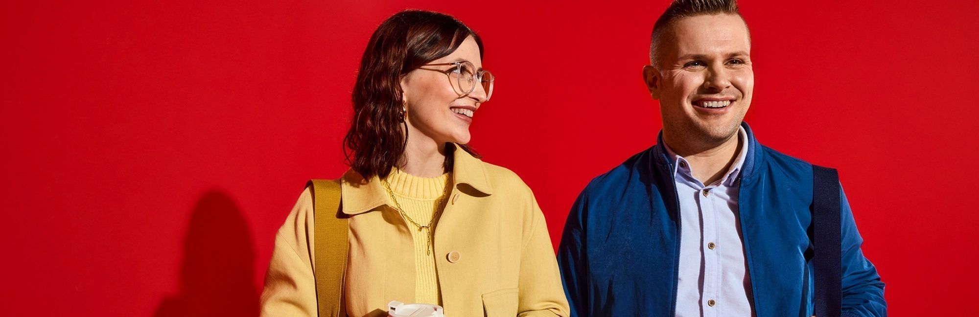 Zwei lächelnde Lidl-Mitarbeiter im modernen Business-Casual-Look posieren gut gelaunt vor einem knallroten Hintergrund.