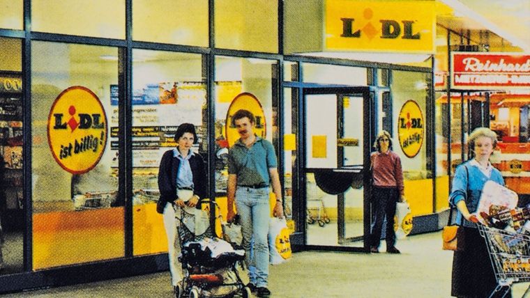 Arhivska fotografija ulaza u retro Lidl trgovinu s kupcima, dječjim kolicima i sloganom 'Lidl ist billig'.