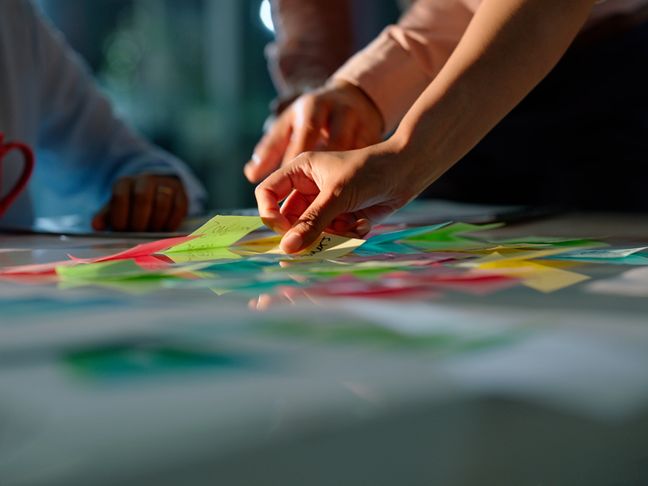 Nahaufnahme mehrerer Hände, die bei Workshop-Atmosphäre farbige Post-its auf einem Tisch neu anordnen.
