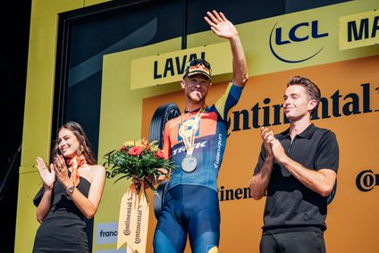 Ein Radrennfahrer im Lidl-Trek-Trikot steht mit einer Medaille und einem Blumenstrauß auf dem Podium und winkt, während zwei Personen neben ihm applaudieren.