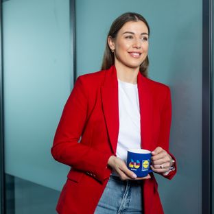 Nasmijana žena u crvenom sakuu drži plavu Lidl šalicu u modernom uredskom prostoru.