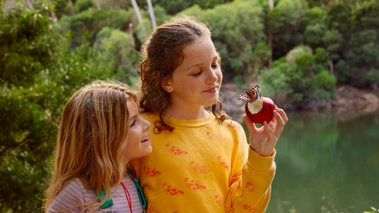 Zwei Mädchen stehen lächelnd am Seeufer, während ein Schmetterling auf dem angebissenen Apfel in der Hand eines Mädchens landet.