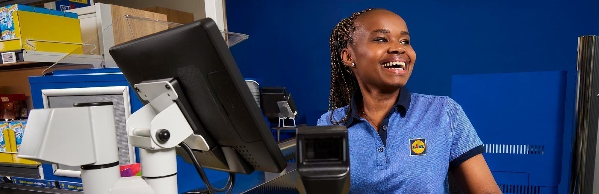 A warmly smiling Lidl cashier scanning a honeydew melon, embodying the fast and friendly service at Lidl checkouts.