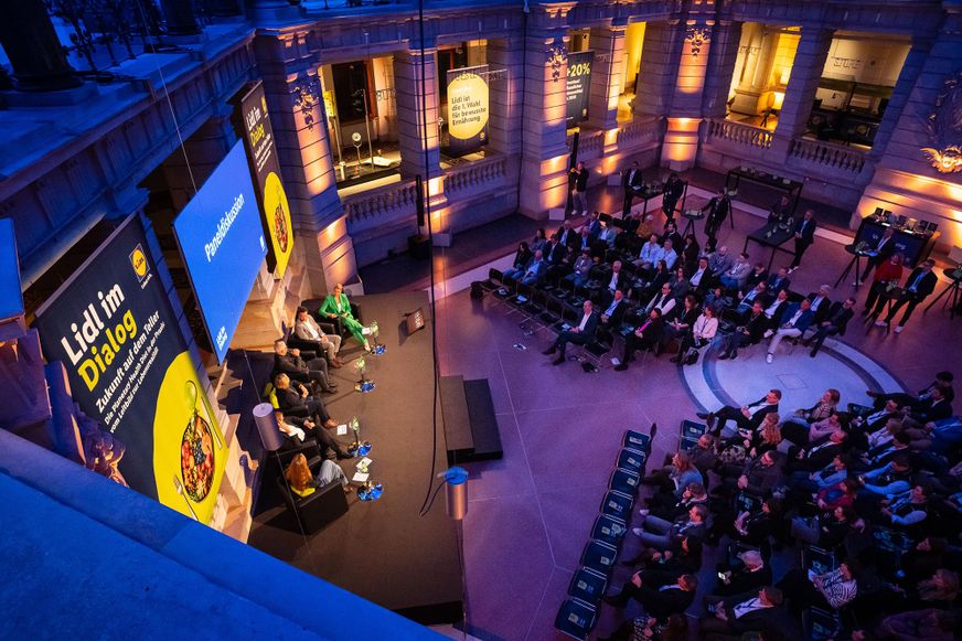 Pogled odozgor na konferencijsku dvoranu s publikom i pozornicom tijekom poslovnog događaja i panel rasprave 'Lidl im Dialog'.