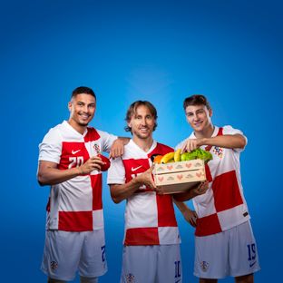 Hrvatski nogometni reprezentativci u dresovima drže drvenu gajbicu punu svježeg voća i povrća.