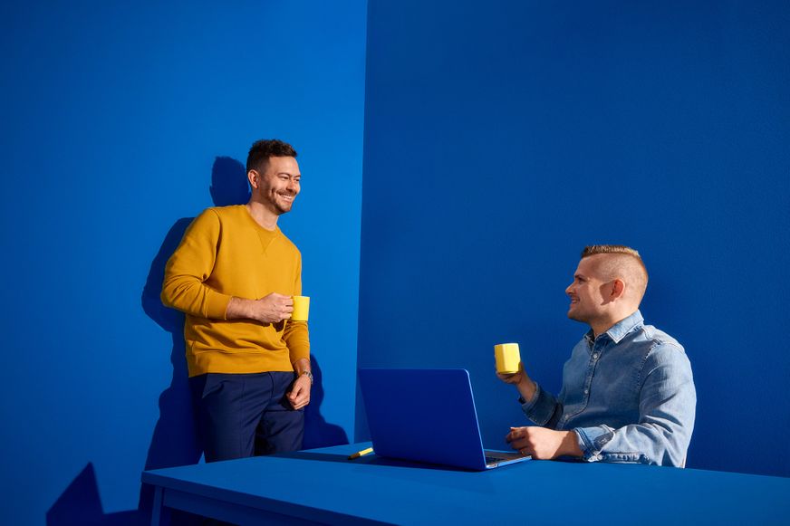 Zwei Lidl Kollegen unterhalten sich gut gelaunt bei einer Tasse Kaffee in einem modern gestalteten, blau-gelben Arbeitsumfeld.
