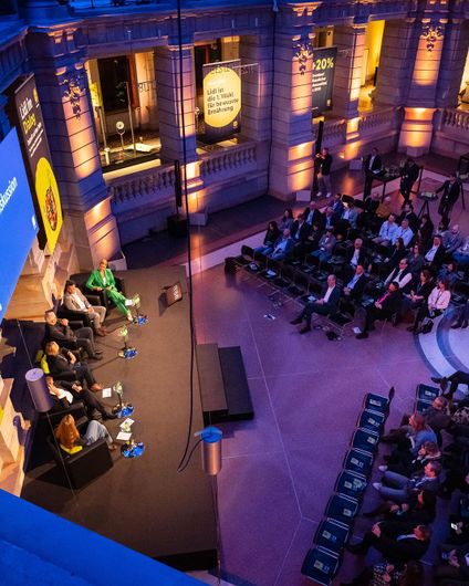 Erhöhte Totale einer Podiumsdiskussion zwischen sechs Experten mit Blick aufs Publikum.
