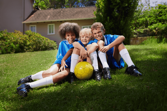 Drei lachende Kinder in blauen Trikots und weißen Stutzen sitzen mit einem gelben Fußball gemeinsam auf einer sonnigen Wiese.