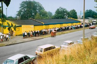 Aufnahme einer langen Menschenschlange vor einer gelben Verkaufshalle mit DDR-Autos im Vordergrund.
