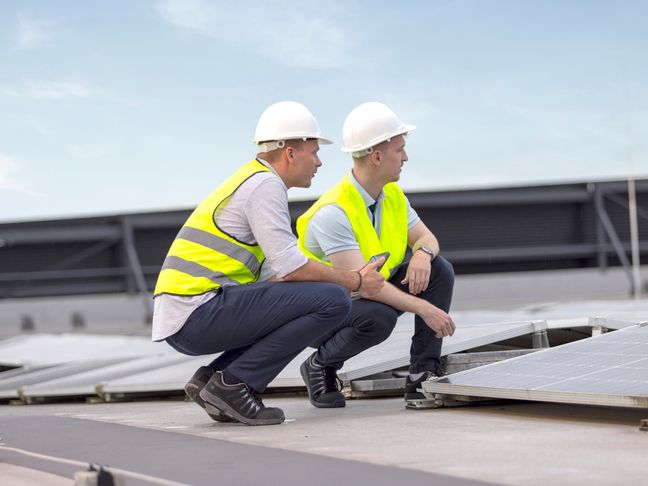 Zwei Fachkräfte hocken in Sicherheitswesten auf einem Flachdach und begutachten eine Photovoltaikanlage.