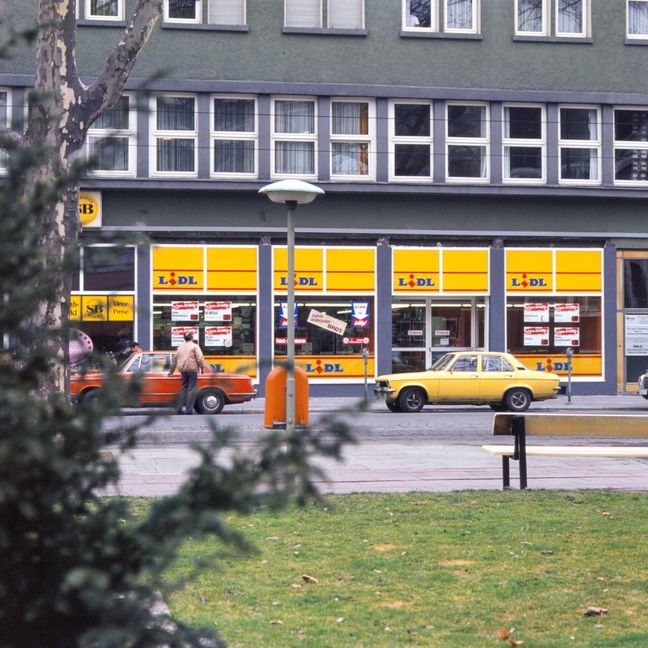Arhivska snimka retro Lidl trgovine u prizemlju zgrade s parkiranim starim automobilima ispred izloga.