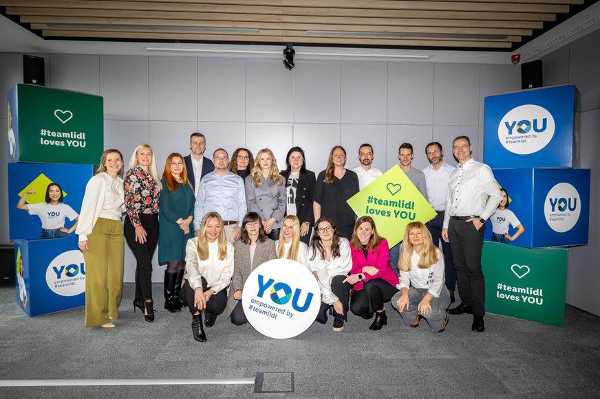 Velika grupa zaposlenika pozira s natpisima i vizualima inicijative 'YOU empowered by #teamlidl' u dvorani.