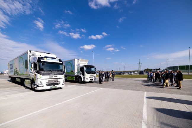 Offizielle Vertreter und Gäste stehen interessiert vor zwei parkenden, vollständig elektrischen Lkws von Lidl.