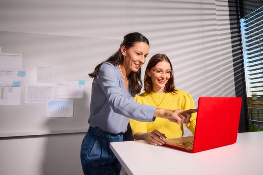 Zwei lächelnde Kolleginnen tauschen sich in einem lichtdurchfluteten Büro gemeinsam über Inhalte auf einem Laptop aus.