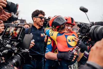 Two cyclists wearing colorful Lidl-Trek jerseys share an emotional hug after a race, surrounded by photographers and camera crews.