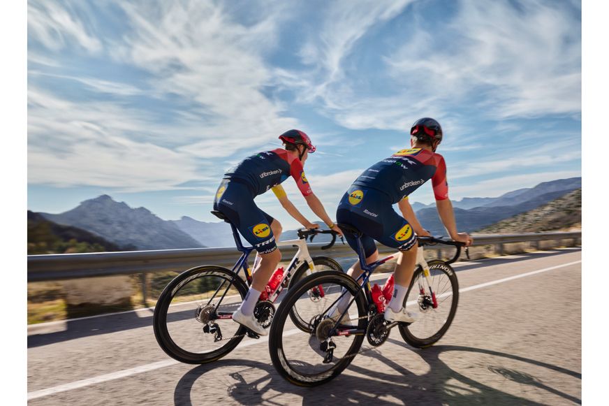 Zwei Radrennfahrer im Lidl-Trek-Trikot fahren nebeneinander auf einer Bergstraße unter einem leicht bewölkten blauen Himmel.