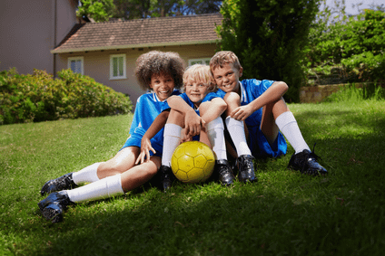 Drei lachende Kinder in blauen Trikots und weißen Stutzen sitzen mit einem gelben Fußball gemeinsam auf einer sonnigen Wiese.