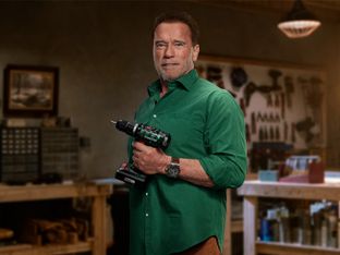Arnold Schwarzenegger hält eine Parkside Akku-Bohrmaschine entschlossen in einer Werkstatt-Umgebung.